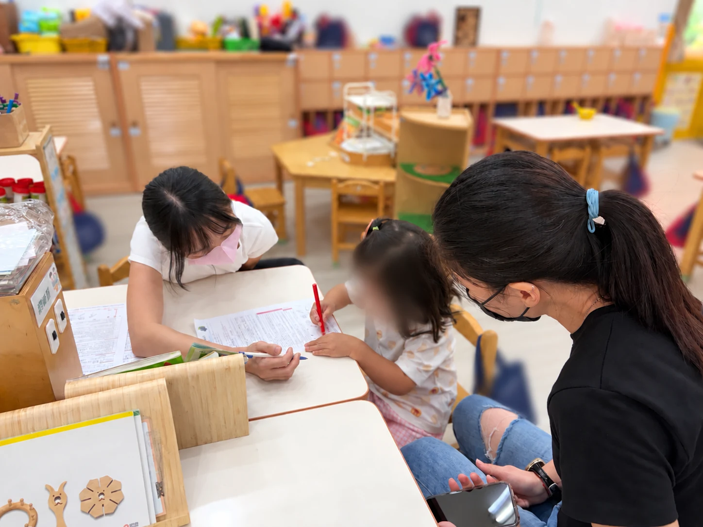 工作人員在桌邊陪幼兒拿筆嘗試，另一位成人在旁觀看